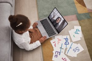 A young girl participates in an online learning session with alphabet cards and a laptop.